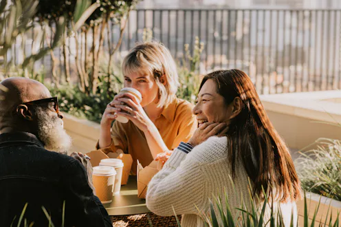 Three colleagues sit outside on a sunny day and enjoy a takeaway lunch and some hot drinks. They are...