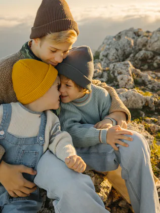 Three children share a tender moment on a rocky hillside at sunset, wearing warm sweaters and beanie...