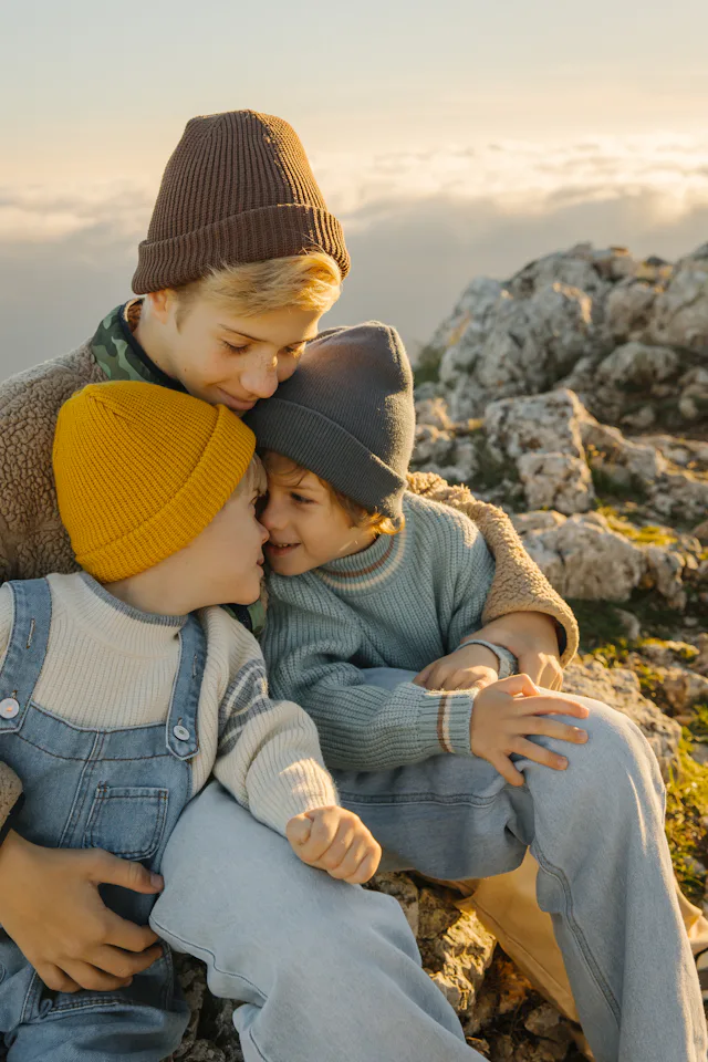 Three children share a tender moment on a rocky hillside at sunset, wearing warm sweaters and beanie...