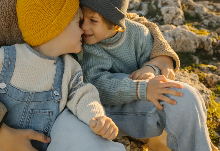 Three children share a tender moment on a rocky hillside at sunset, wearing warm sweaters and beanie...