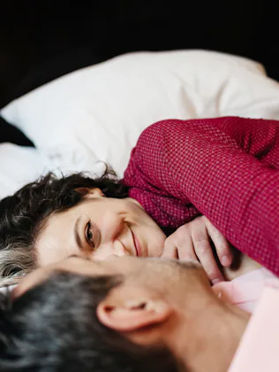 A mature, loving couple is looking at each other lying on a hotel bed. They seem to be in love as th...