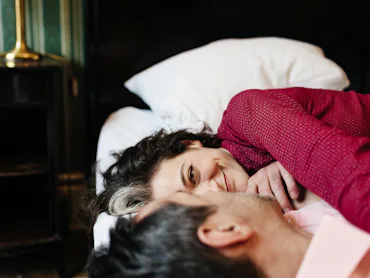 A mature, loving couple is looking at each other lying on a hotel bed. They seem to be in love as th...
