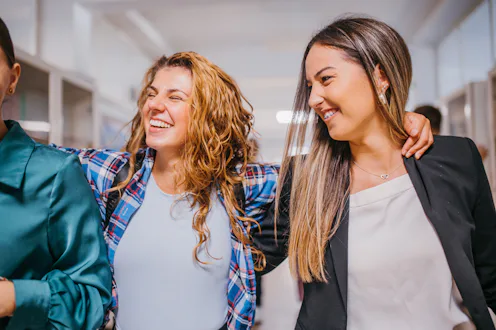 Three diverse young women are walking arm-in-arm down a hallway, laughing and sharing a moment of jo...