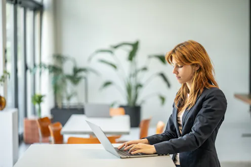 Young adult woman typing on a laptop at a white desk in a corporate office