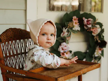 An adorable baby girl, nine months old, sitting in a high chair on the porch of a charming cottage. ...