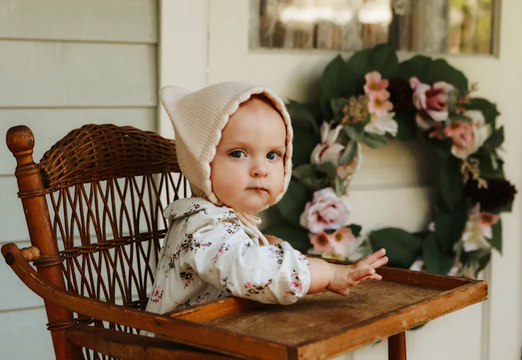 An adorable baby girl, nine months old, sitting in a high chair on the porch of a charming cottage. ...