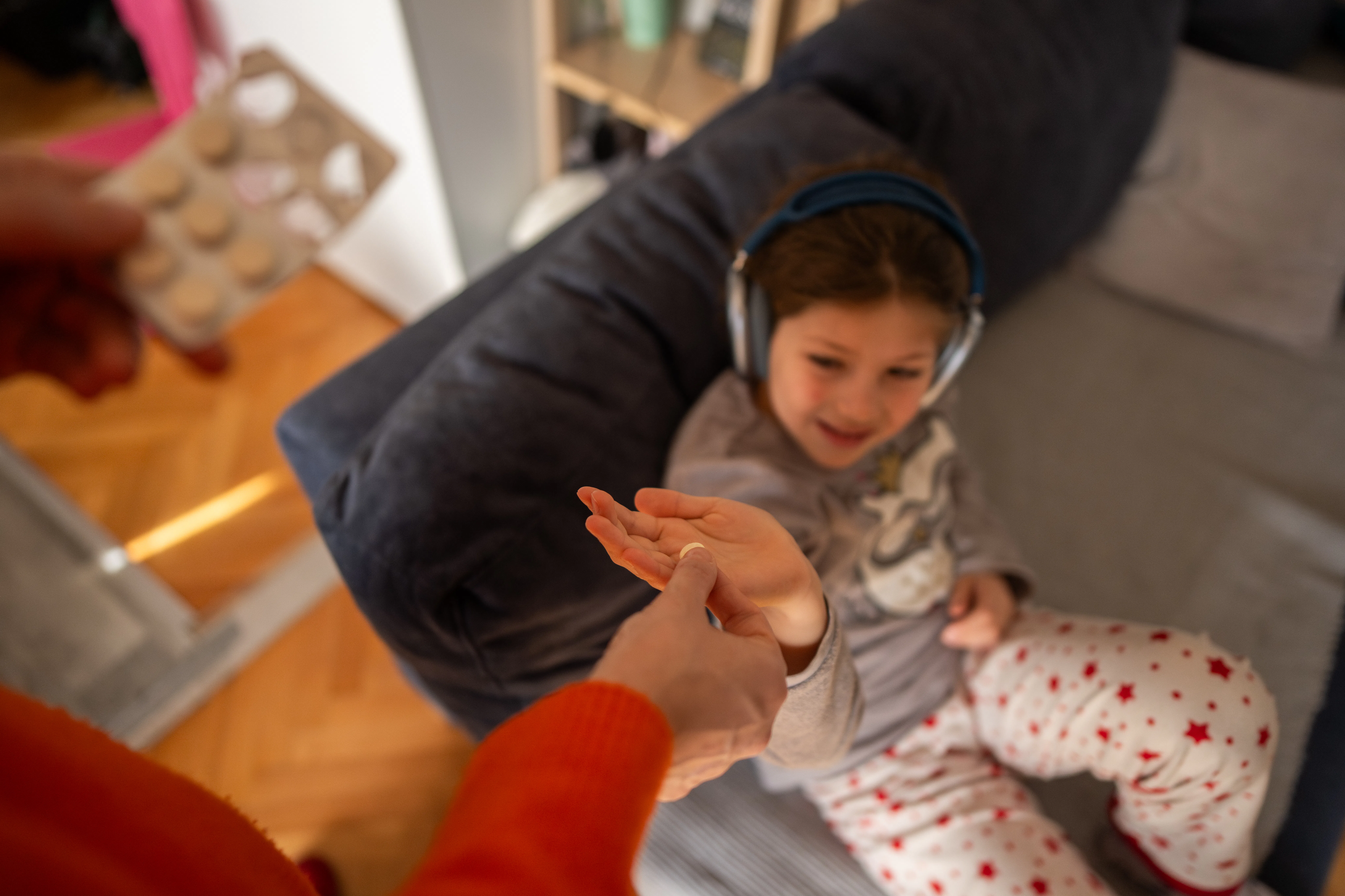 A parent hands a child a pill on a living room sofa as the relaxed child listens to headphones; inti...