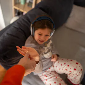 A parent hands a child a pill on a living room sofa as the relaxed child listens to headphones; inti...