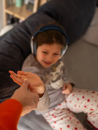 A parent hands a child a pill on a living room sofa as the relaxed child listens to headphones; inti...