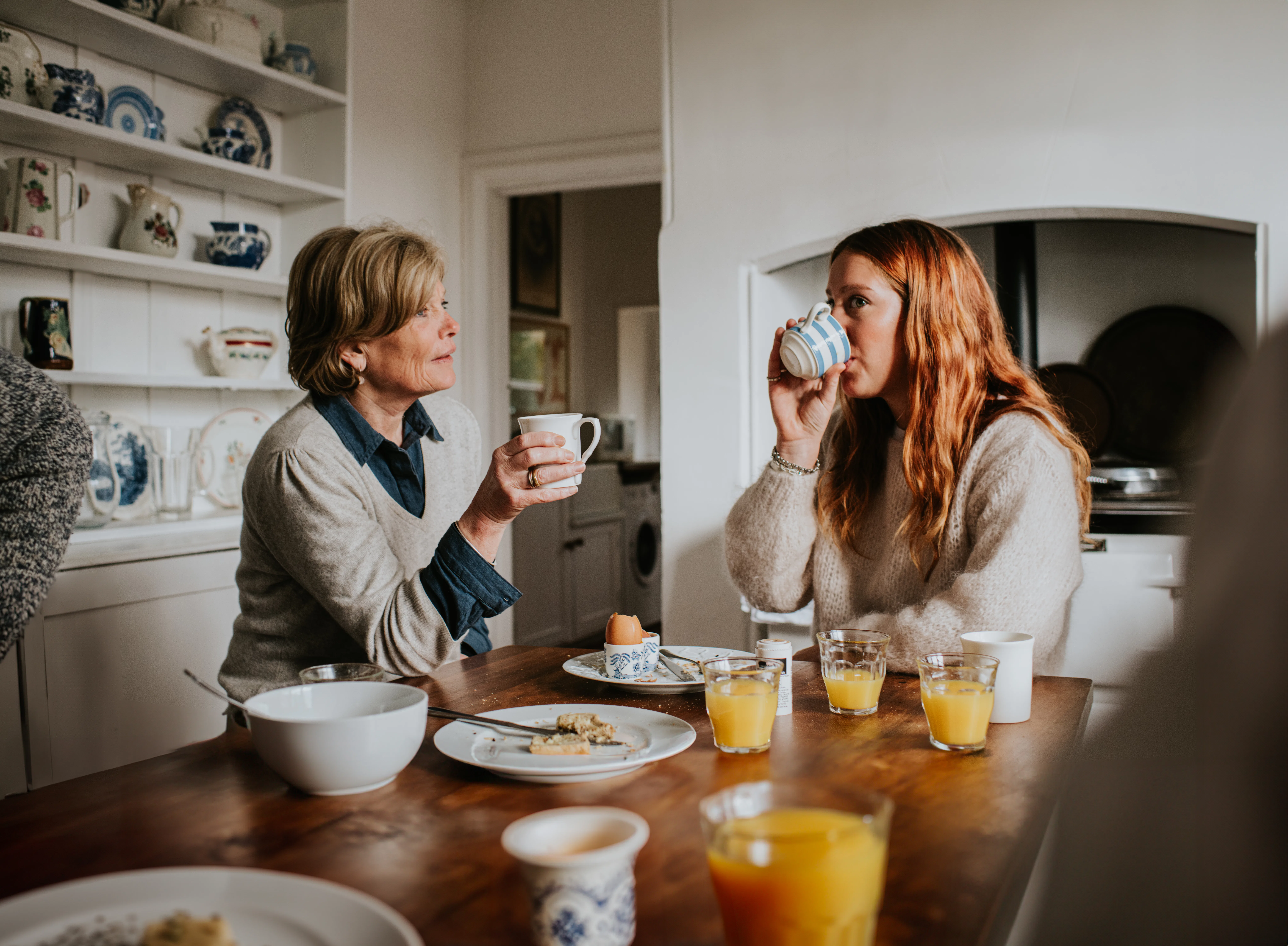 An older woman sits beside a younger woman in a stylish kitchen. They both drink coffee and tea from...