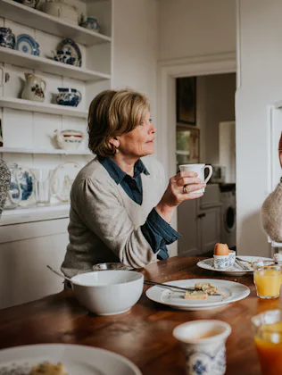 An older woman sits beside a younger woman in a stylish kitchen. They both drink coffee and tea from...