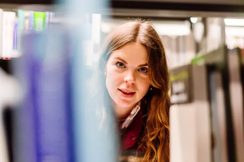 A horizontal close-up shot of a smiling young woman wearing a colorful fleece jacket reading a book ...