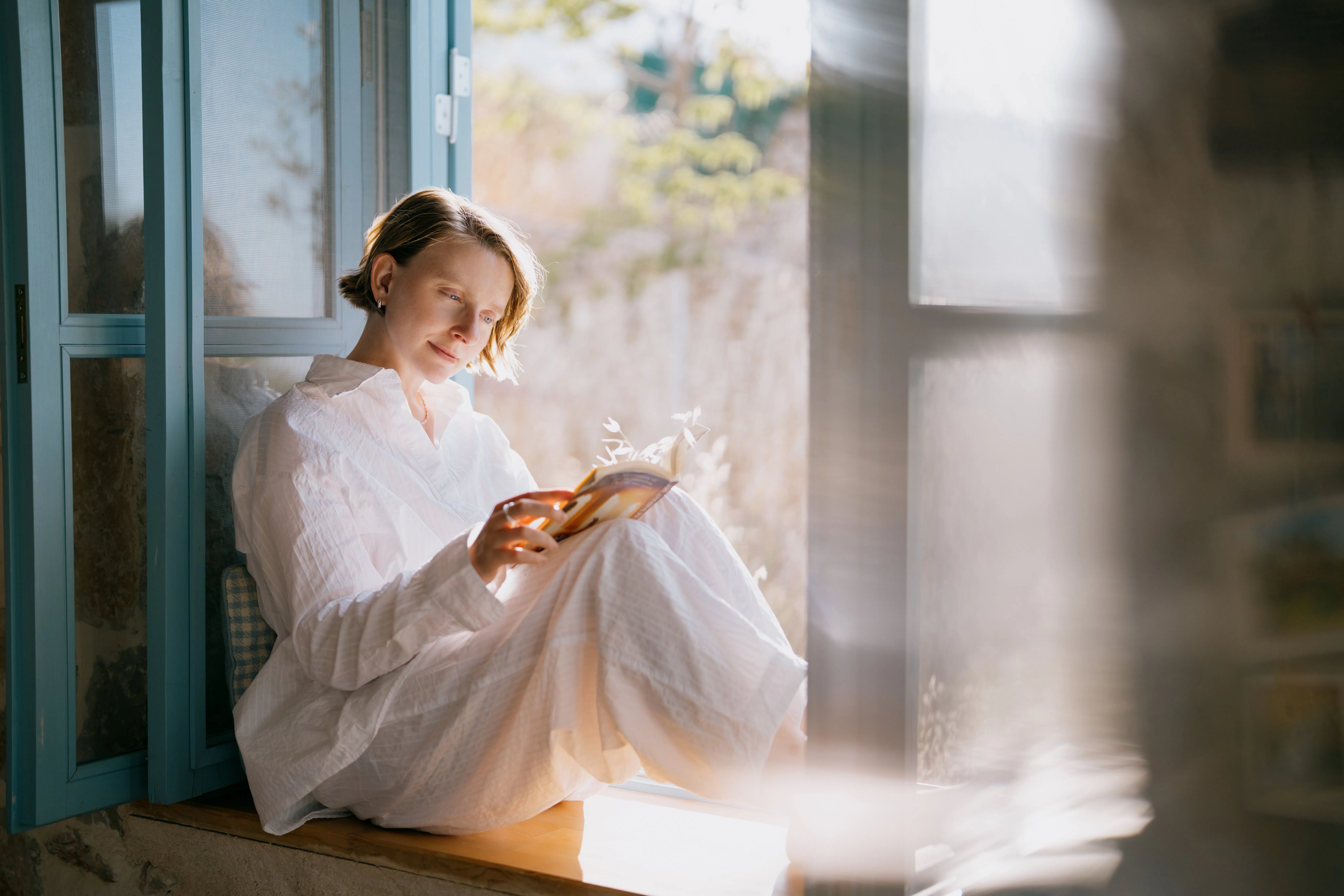 A serene woman sits by a sun-drenched window, engrossed in reading a book. The soft natural light an...