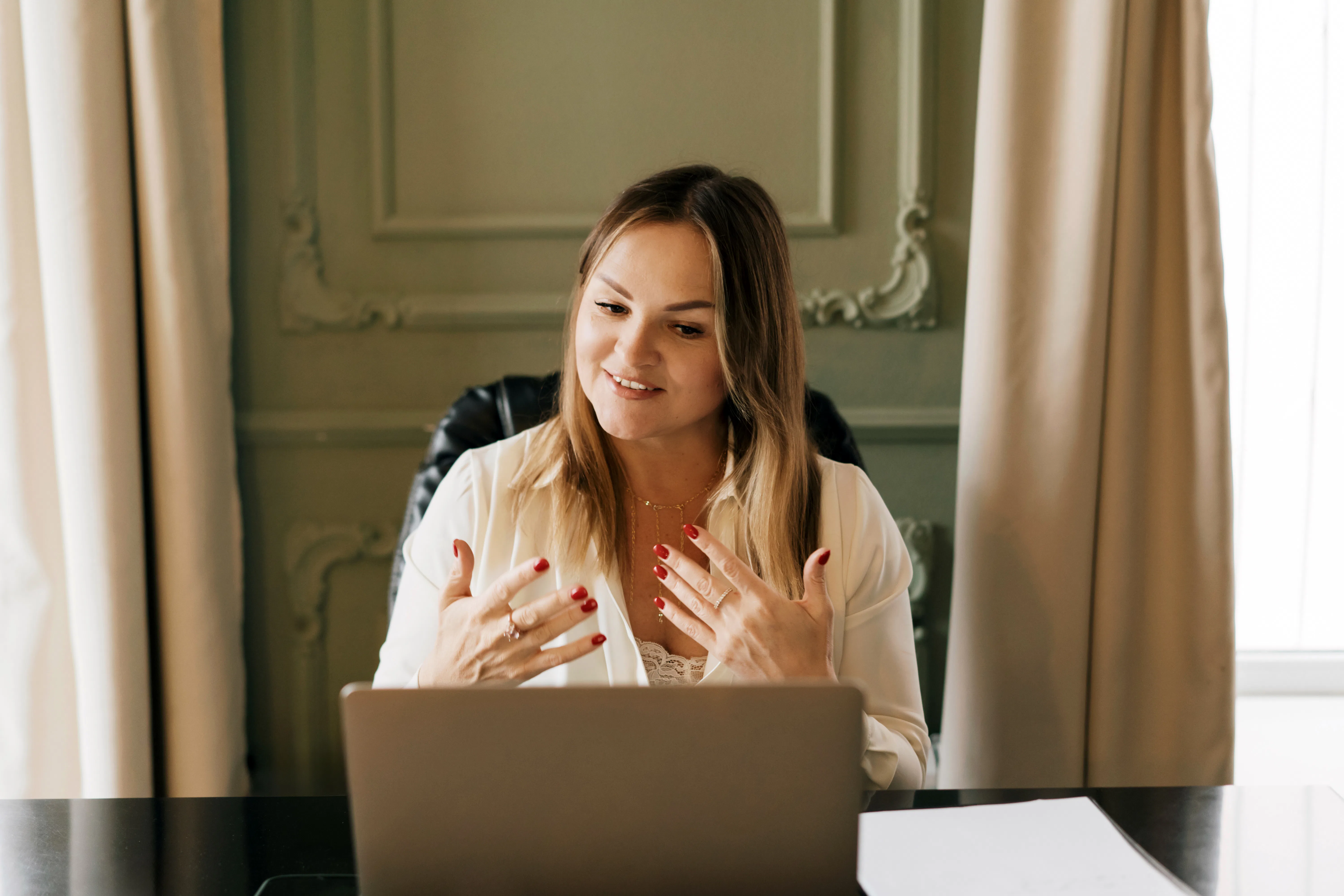 A professional female therapist sits at a desk with a laptop, speaking gently and gesturing during a...