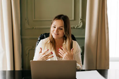 A professional female therapist sits at a desk with a laptop, speaking gently and gesturing during a...
