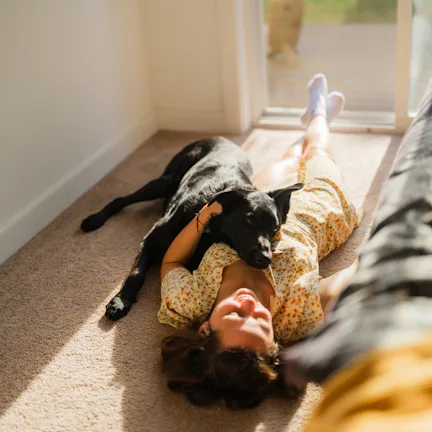 Woman in a summer dress lying on a carpet in a sunlit room near a window, relaxing with her dog. Sof...