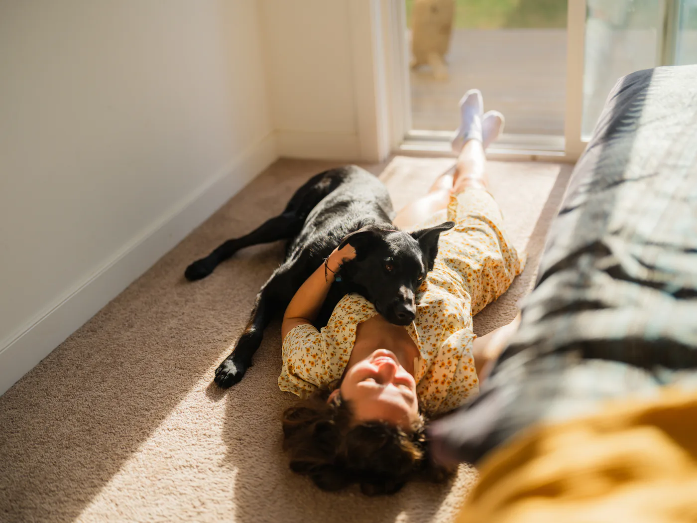 Woman in a summer dress lying on a carpet in a sunlit room near a window, relaxing with her dog. Soft natural light streams through the window, casting warm highlights on the floor. The dog is cuddled up beside her, both appearing calm and content in the peaceful indoor moment.