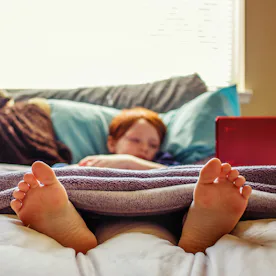 7 year old boy watching videos on his laptop while lying in bed -- image taken indoors using natural...