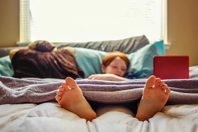 7 year old boy watching videos on his laptop while lying in bed -- image taken indoors using natural...