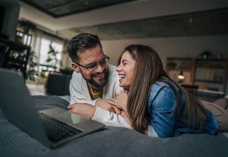 A couple is lying side by side on a large bed. They look at the screen of laptop relaxed and satisfi...