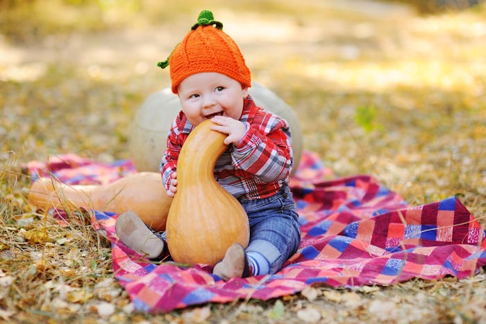A baby with a pumpkin knit beanie biting in a large pumpkin while sitting on a blanket in a park