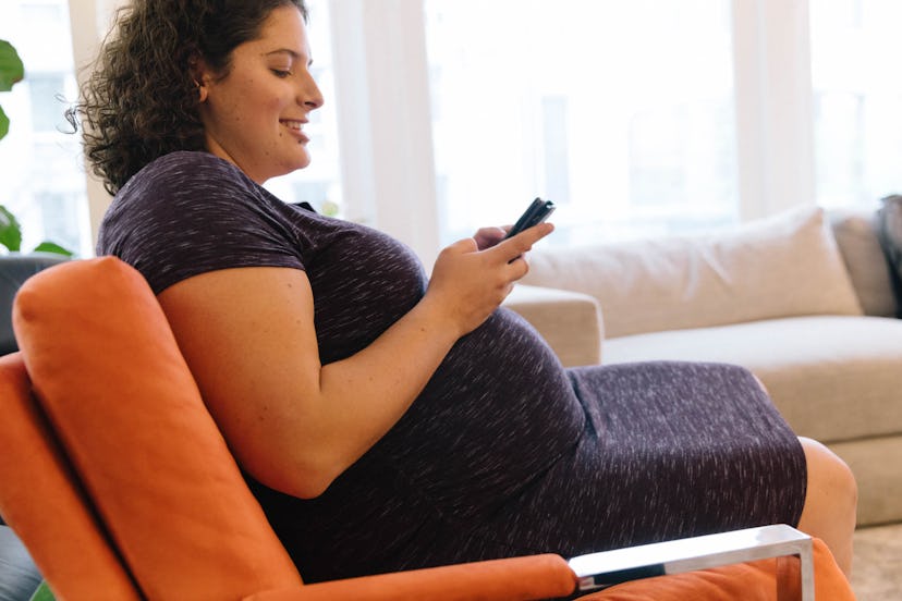 A pregnant woman in a black dress is sitting on the couch and looking at her mobile phone