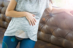 A pregnant woman in a grey t-shirt and blue jeans leaning on a couch while holding her belly