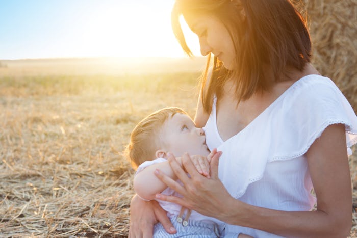 A mother in a white top breastfeeding her baby outside with the sunset behind them