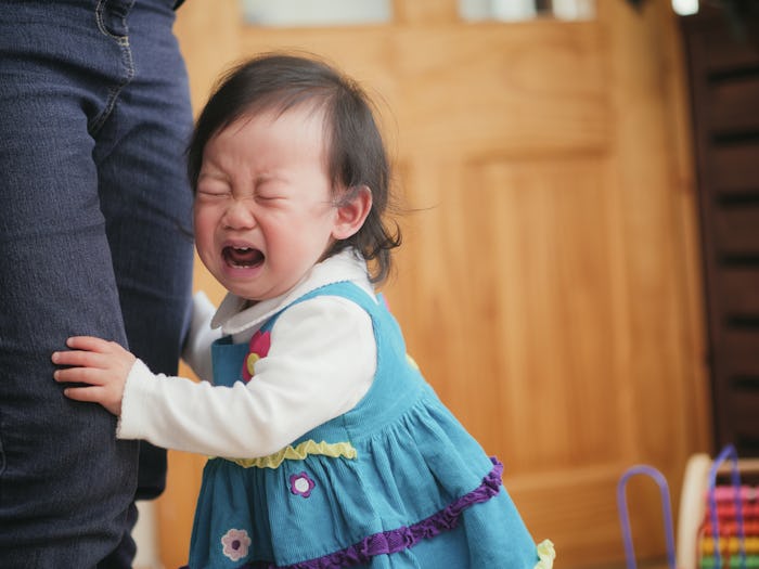 A little daughter in a dress crying during a tantrum in public holding on to her mom's legs