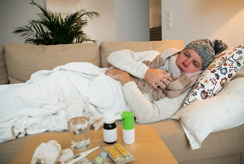 man huddles on couch in beanie, blanket, and scarf