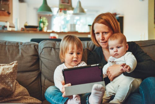 A work-at-home mom sitting on a couch holding her baby and her toddler while they're looking at a ta...