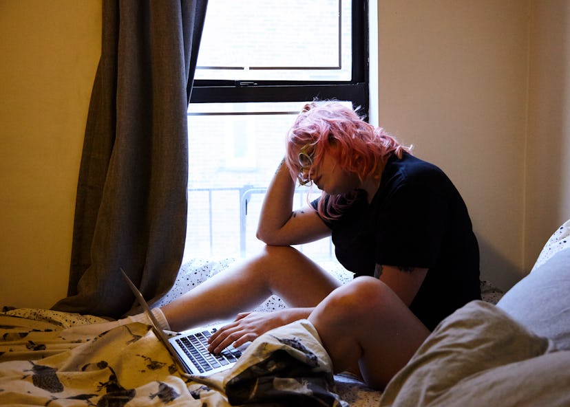 Woman sitting on the bed in her room with the laptop in front of her, typing.