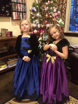 Two girls standing in front of a Christmas tree, one wearing a blue, and the other one wearing a pur...