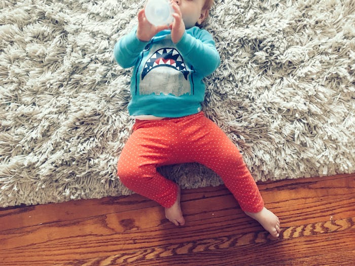 A toddler lying on a shag carpet wearing a blue shirt with a shark and orange leggings drinking milk