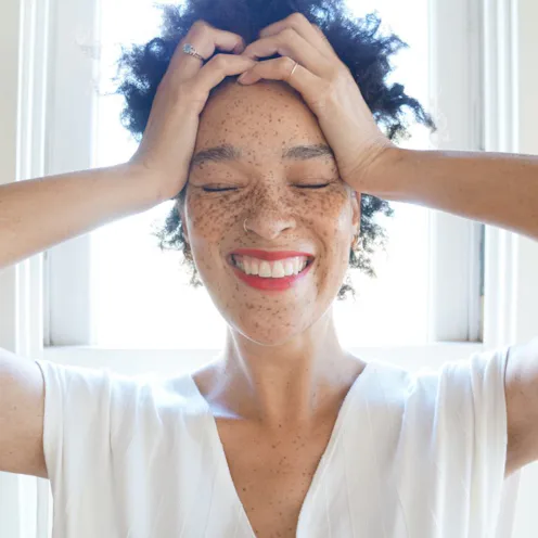 A woman with short curly hair holding her head and smiling with her eyes shut.