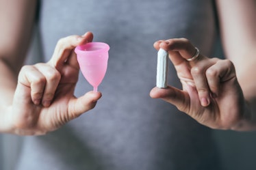 A woman in a grey shirt holding a pink menstrual cup, and a tampon