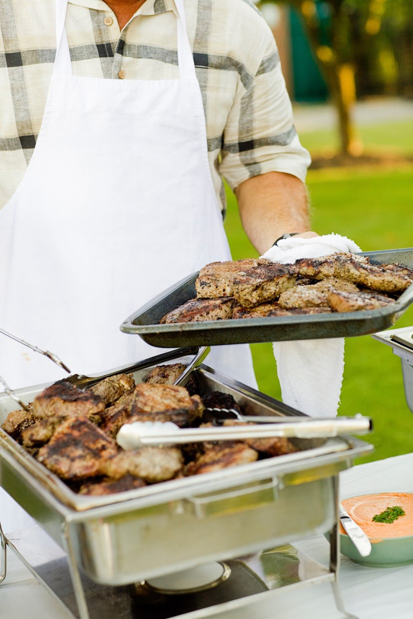 A man wearing a white apron holding a plate of meat while barbecuing