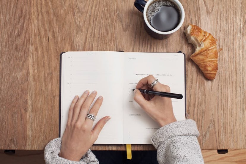 A woman wrriting down budget plans in a notebook, with a croissant and coffee
