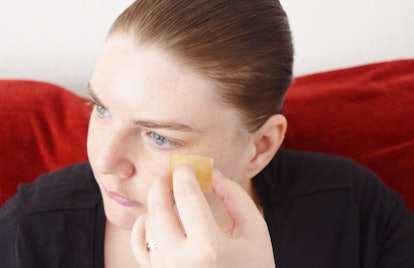 A woman putting a green ice tea cube under her left eye 