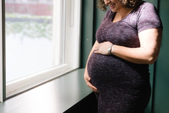 A pregnant woman holding her stomach next to a window, wondering if her pregnancy weight gain affect...