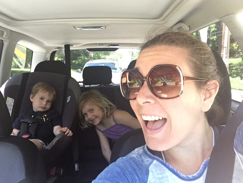 A woman taking a selfie in the car with her kids in the back seat smiling.