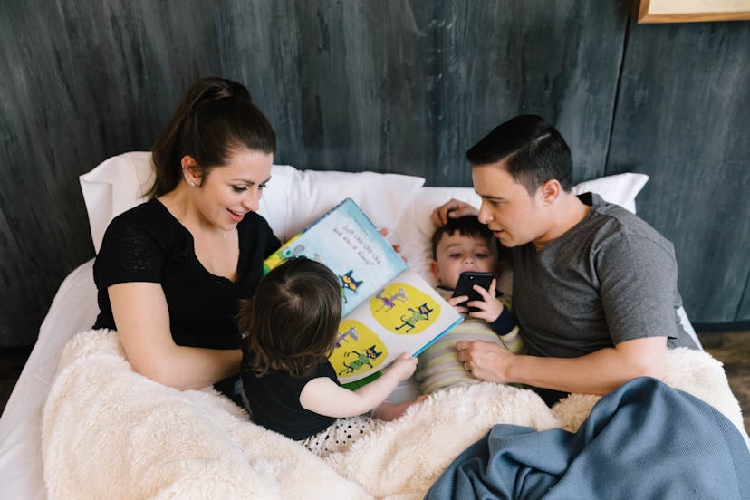 Parents lying in bed next to their kids while a girl reads a book and a boy looks at a phone.