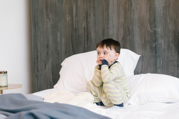 A toddler boy in his beige-navy pajamas squatting on a bed, experiencing teething