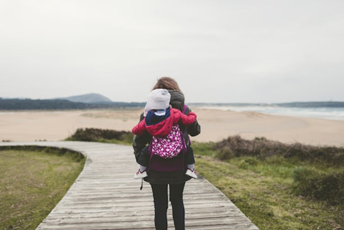 Single mom carrying her kid piggyback down a wooden path towards the sea