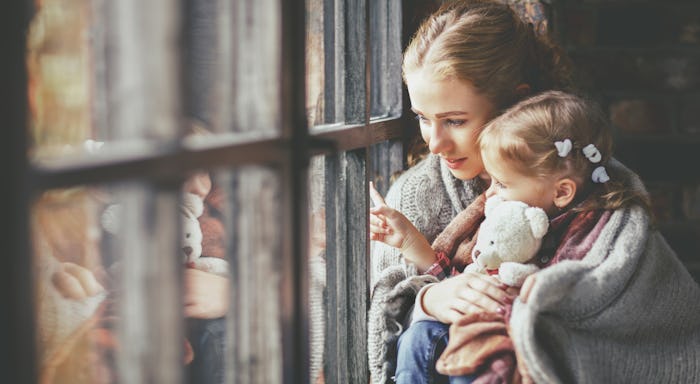 A feminist mom looking out of a window with her toddler in her hands both covered by a blanket