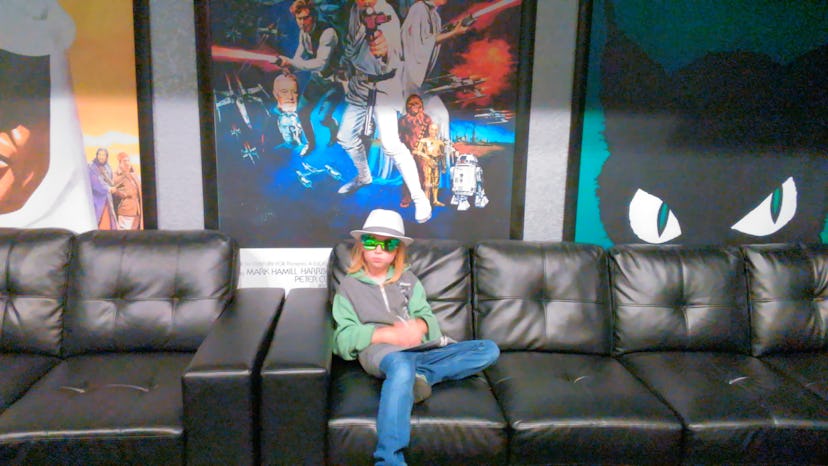 A young kid sitting on a couch with posters behind while wearing a hat and sunglasses
