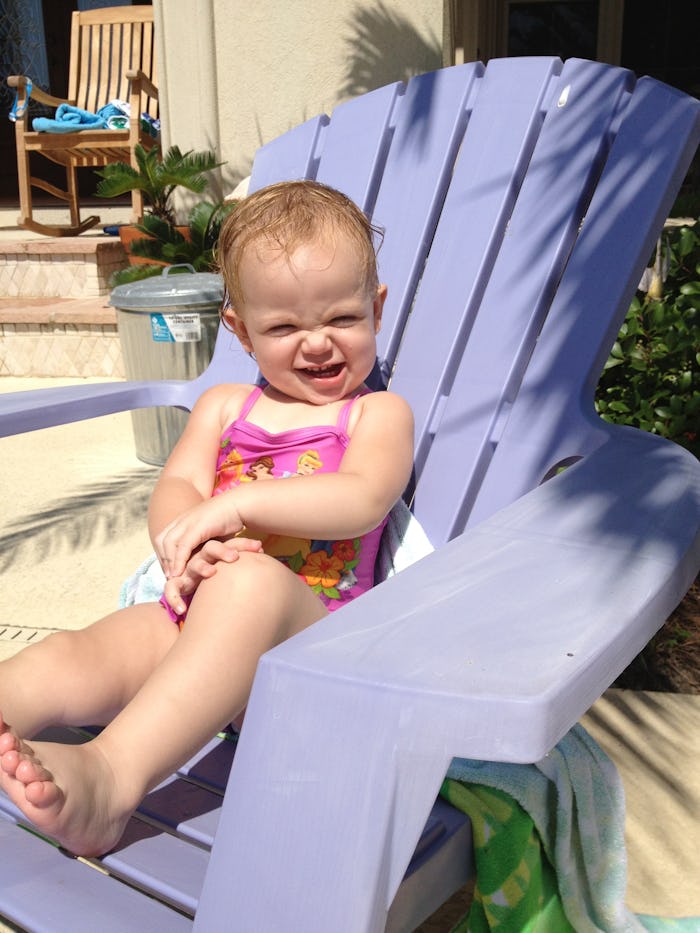 A toddler girl in a pink bathing suit sitting on a periwinkle lounge chair outside and smiling