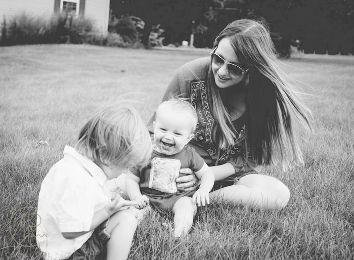 An anxious mom sitting on grass and smiling with her baby and her toddler