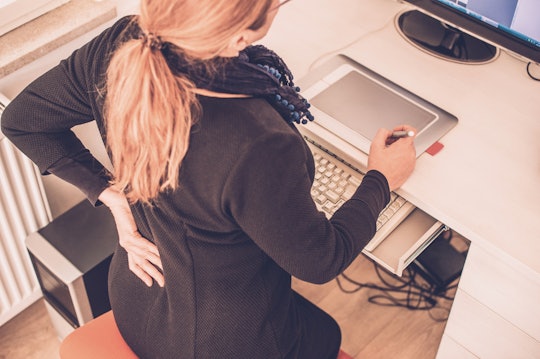 A woman sitting on a high chair working on her computer while holding onto her back as it is in pain...