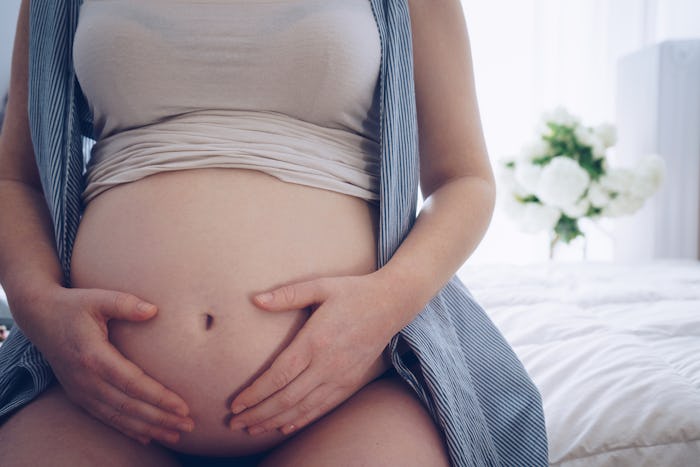 A hormonal pregnant woman sitting on her bed with her hands on her exposed belly